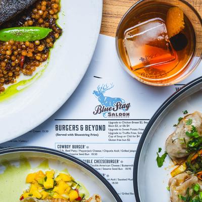Multiple food plates with Old Fashioned and a menu on a wooden table.