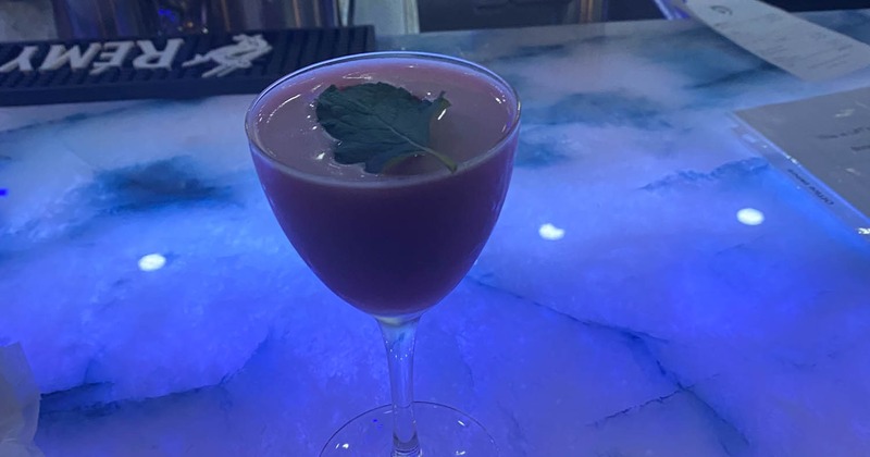 A red cocktail garnished with a mint leaf, resting on a bar top