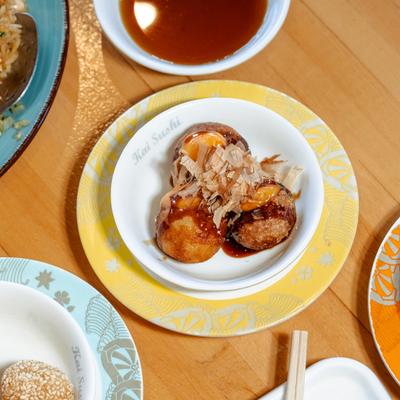 Takoyaki on white plate over yellow saucer, surrounded by other dishe.