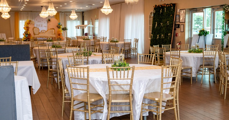 A banquet hall with round tables covered in white cloths and surrounded by gold chairs, decorated for an event