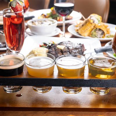 Beer flight with cocktails and plates of food in the background.