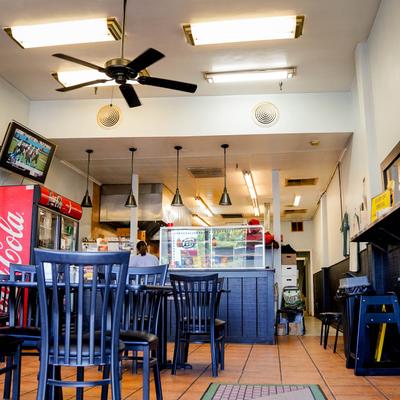 Pizzeria interior with empty black chairs and tables, a counter, TV, and warm lighting.