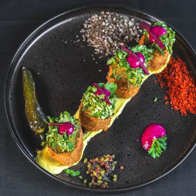 Black plate with five garnished croquettes, sauces and colorful garnishes.