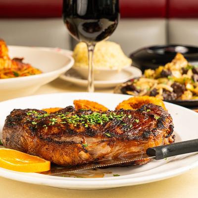 Ribeye steak on a white plate with onion rings, a glass of red wine and food is in the background.