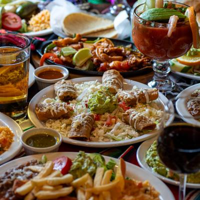 Mexican tables spread with fried tacos, shrimp cocktail, fajitas, guacamole, and drinks.