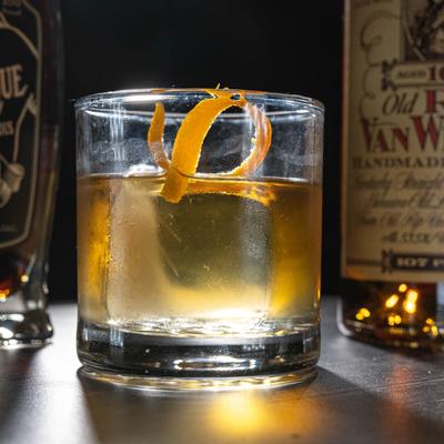 Bourbon cocktail with a spiral orange peel garnish is centered between bourbon bottles.