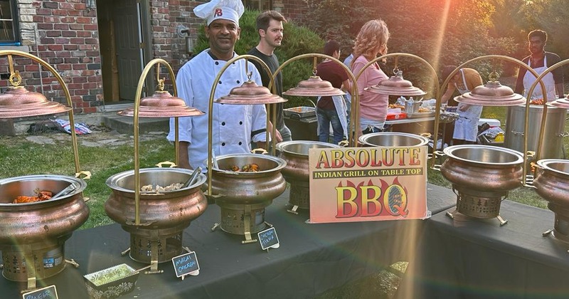 Exterior, a cook standing by a buffet table with chafing dishes filled with Indian grill food