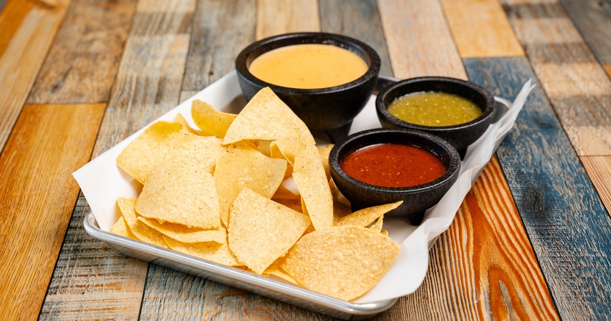 Tortilla chips served on a tray with a trio of dips in black stone bowls