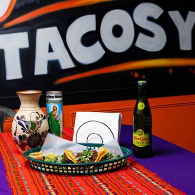 Carne asada tacos on a table beside a drink, a vase, and a devotional candle.