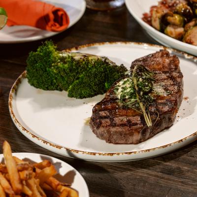 A grilled ribeye steak sits on a plate with broccoli, surrounded by other food plates.