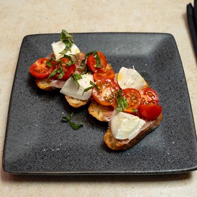 Toasted bread topped with mozzarella, cherry tomatoes, prosciutto, and fresh basil garnish.