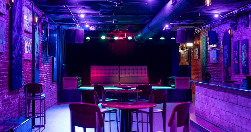 Dimly lit nightclub interior with purple and blue lighting, small stage, and scattered tables and chairs