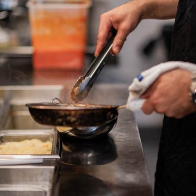 Chef using metal tongs to handle food inside a metallic skillet on a stove.