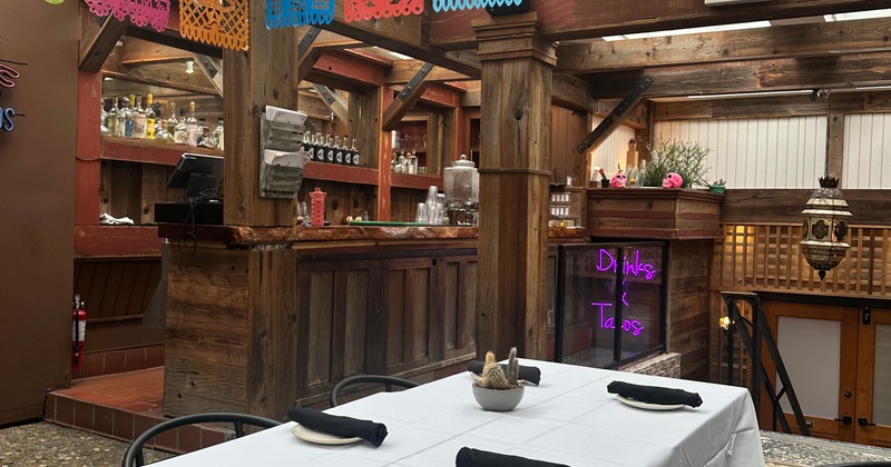 Rustic interior with a wooden bar, papel picado banners, and a neon sign reading Drinks & Tacos