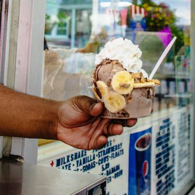 A hand holding a dessert cup filled with chocolate ice cream, banana slices, and whipped cream.