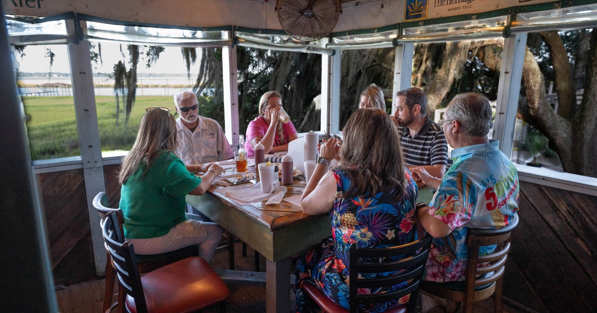 A group of people sit around a table outdoors, with a grassy landscape and trees in the background