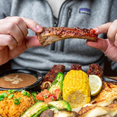 A person holding a barbecue rib above a platter of rice, beans, and corn.