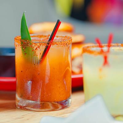 Two cocktails with chili-salt rims and red straws on a table.