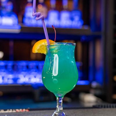 Blue cocktail with an orange slice and a cherry on a bar counter.