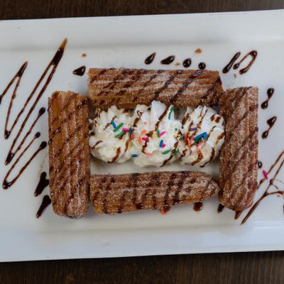 Churros with whipped cream, rainbow sprinkles, and chocolate drizzle.
