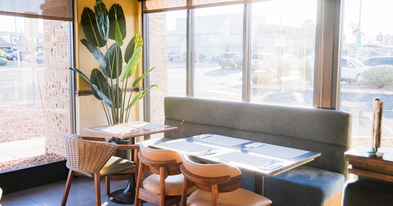 Sunlit restaurant interior corner with large windows, a green plant and seating around a set table