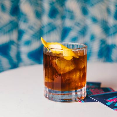 A glass of dark cocktail with ice and a lemon twist on a white table.