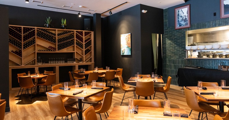 Interior with wooden tables, tan leather chairs, a large wine rack, a painting, and an open kitchen