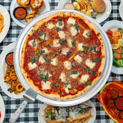 Large margherita pizza surrounded by Italian dishes on checkered tablecloth.