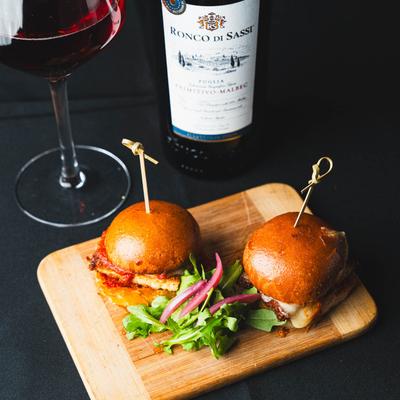 Two gourmet sliders on a wooden board with arugula salad, served with red wine.