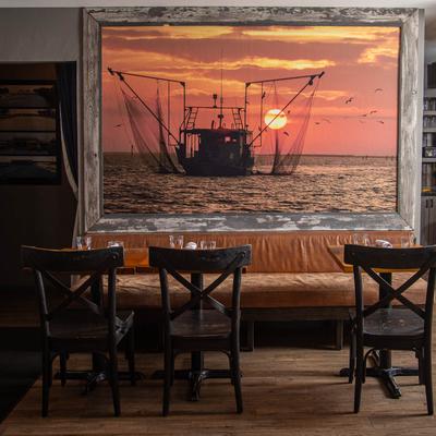 Seating with tables and chairs by a large framed wall poster of a boat at sunset.