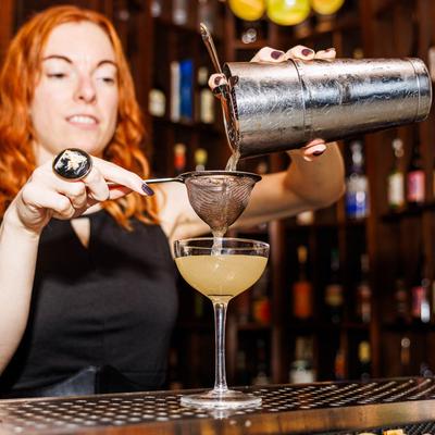 A bartender pours a cocktail through a strainer into a glass.