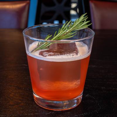 A glass of Solstice Smash, with a large ice cube and a rosemary sprig.