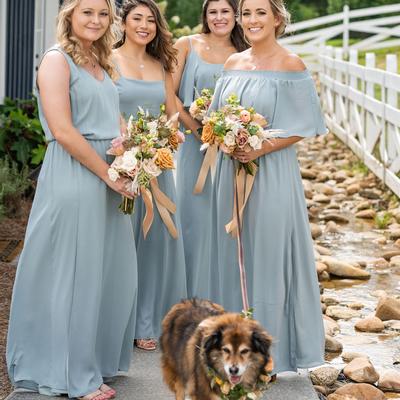 Four smiling bridesmaids stand on a path beside a small stream holding floral bouquets