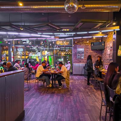 A lively restaurant scene with guests enjoying meals, vibrant decor, and colorful lights.