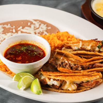 Birria tacos on a plate with rice, refried beans, consome, and lime.