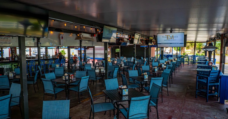 Outdoor covered patio with numerous blue chairs and tables, with TVs mounted overhead