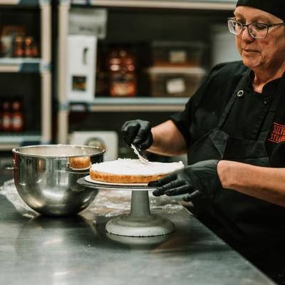 Person frosting a cake on a metal stand in the kitchen.