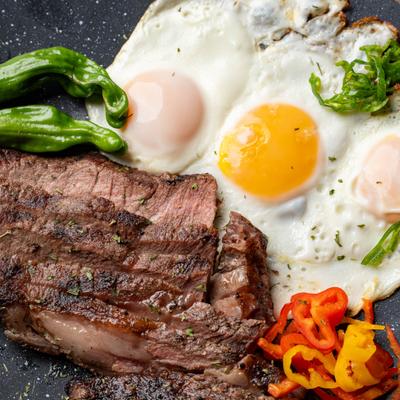 Sliced steak served with fried eggs, two green peppers, sliced red and yellow peppers.