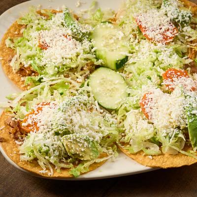 Four crispy corn tortillas topped with shredded chicken, lettuce, avocado, and cheese.