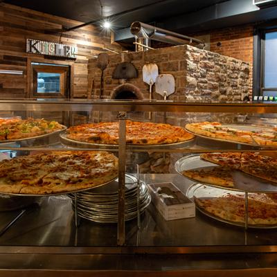 Display case with several pizzas and stone wood-fired oven in the background.