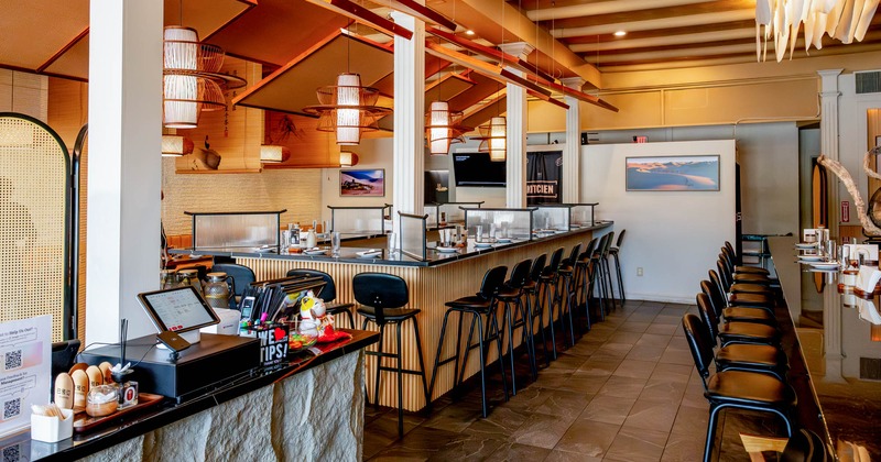 Interior of a modern restaurant with a sushi bar, featuring pendant lights, a bar with black stools