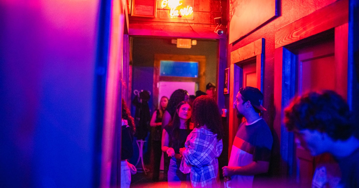 Dimly lit hallway with a neon sign, featuring people engaged in conversation