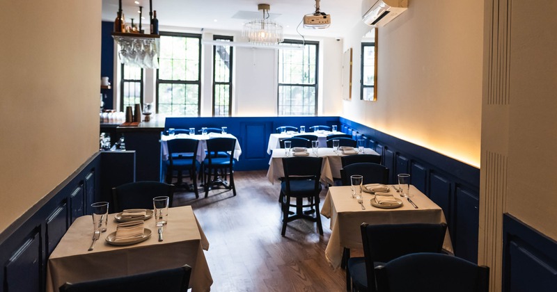 A small dining room with white tablecloth-covered tables, blue and white walls, and large windows