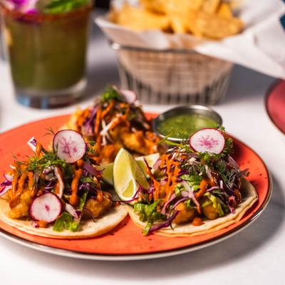 Three shrimp tacos on a red plate are topped with radish slices, greens, and sauce.