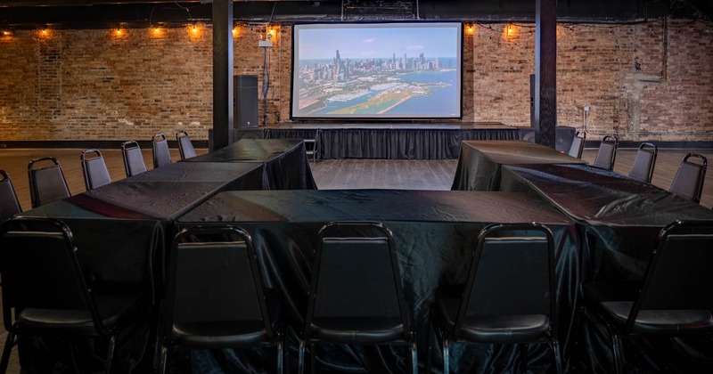 Event venue with a large projector screen, brick walls, and black tables