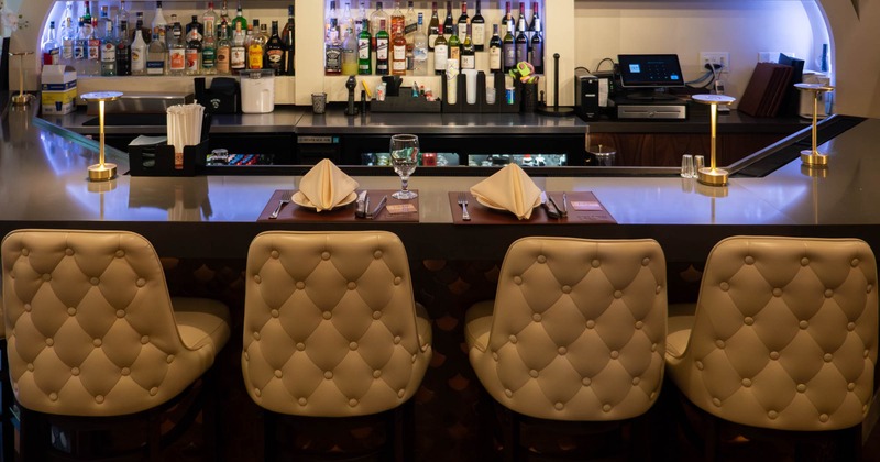 A stylish bar with beige tufted leather stools, a polished counter, liquor bottles on shelves