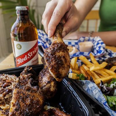 A hand holds a jerk chicken drumstick over the dish plate, a beer bottle and salad are nearby.
