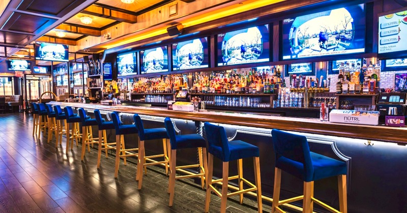 The bar area with stools, bottles on the shelves, and TVs playing sports