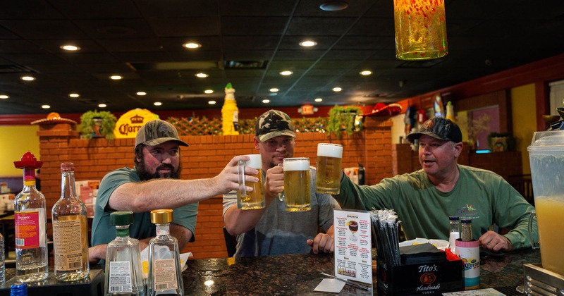 People toasting with beer at a bar with liquor bottles in the foreground