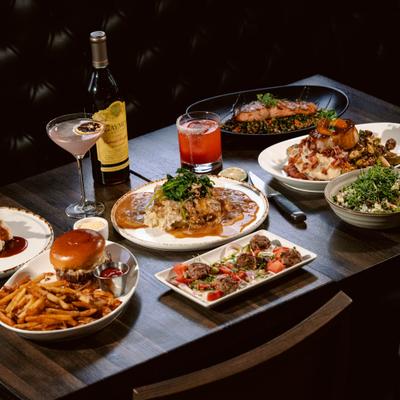 A dark wood table filled with various gourmet dishes, cocktails, and a wine bottle.